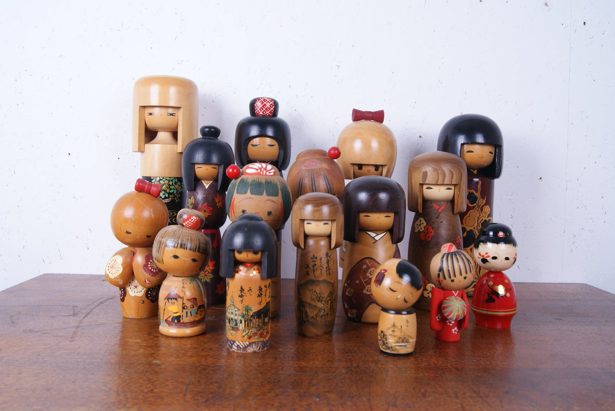 A collection of traditional Japanese kokeshu wooden doll figures in various sizes and designs, displayed on a wooden table against a plain white wall.
