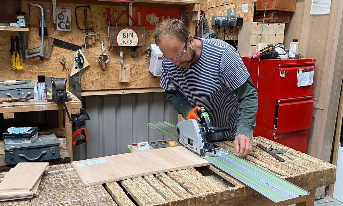 A man wearing safety glasses operates a circular saw guide rail system in a woodworking workshop, cutting a light-colored wood board. The workshop is filled with tools, wood panels, and a red tool chest in the background.