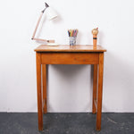 A vintage wooden writing desk with a single drawer, shown against a white wall with a black floor. On top of the desk are a white desk lamp, a cup of colored pencils, and a small figurine.
