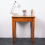 A vintage wooden writing desk with a single drawer, shown against a white wall with a black floor. On top of the desk are a white desk lamp, a cup of colored pencils, and a small figurine.