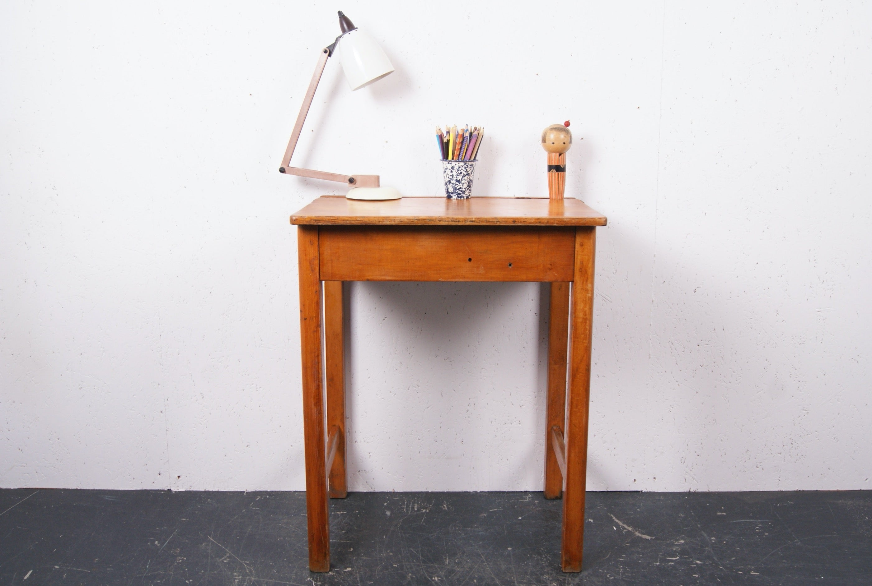 A vintage wooden writing desk with a single drawer, shown against a white wall with a black floor. On top of the desk are a white desk lamp, a cup of colored pencils, and a small figurine.