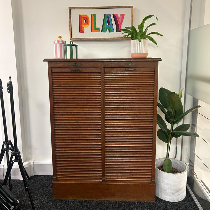 A mid-century modern wooden cabinet with two hinged doors, styled in a room setting with a potted plant, decorative items, and a framed 'PLAY' sign on a white wall.